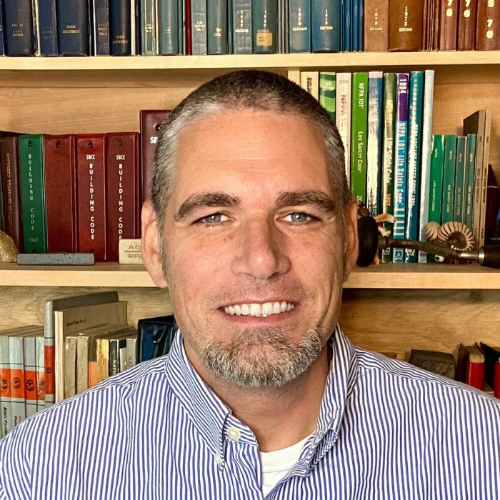 Headshot of Glenn Mathewson in a striped shirt in front of a bookcase