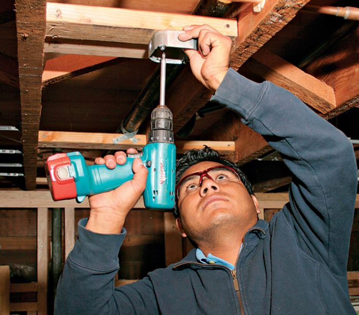 Man using a power driver to attach a metal box to blocking between joists.