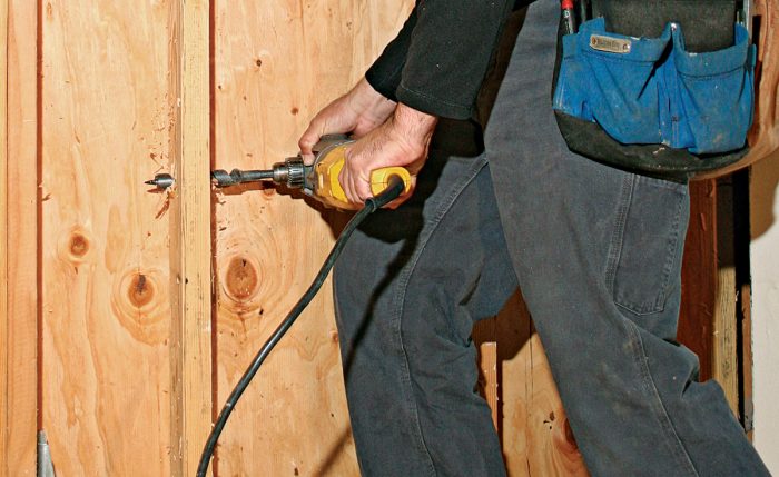 Man using a power drill rested on his thigh to make a hole in a stud to pull a cable through.