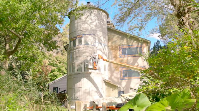 Wide shot of silo attached to house with a painter spray painting the siding