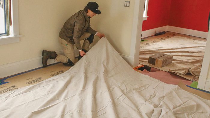 Photo of man in a brown jacket and black hat covering floors with tarp.