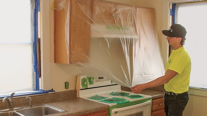 Photo of a man in a neon yellow shirt covering appliance surfaces with plastics and tape.