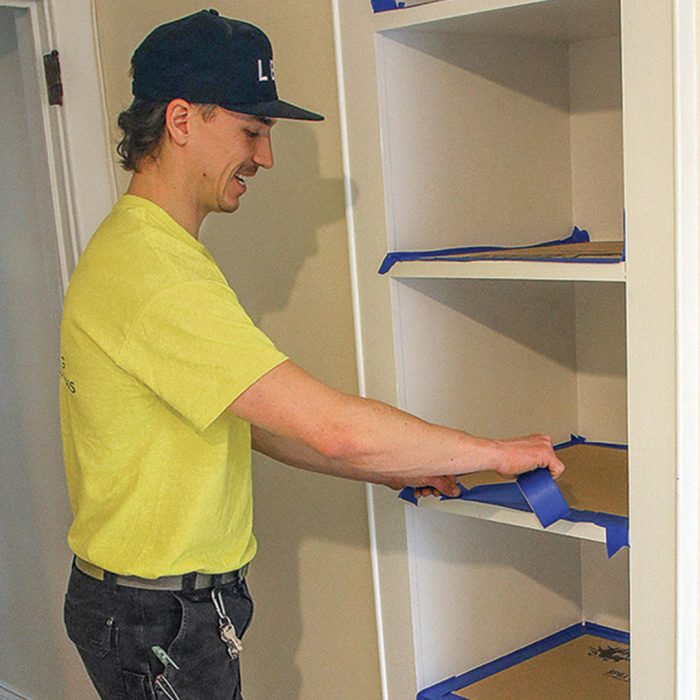 Photo of a man in a neon yellow shirt putting masking tape on shelves and cabinets.