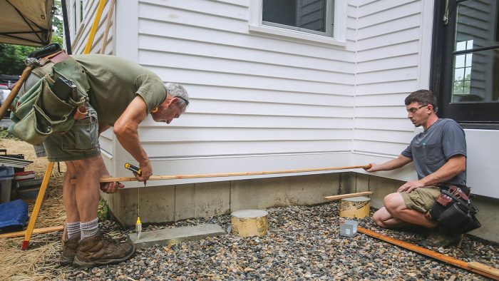 Two men holding a deck board as they measure and mark the length of a wall.