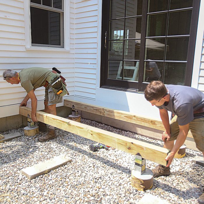 Two men pushing a beam braced in two post bases towards the side of a house. 